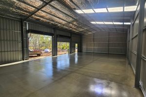 Large infill shed slab poured inside an erected portal-frame kit shed, bush setting visible through the roller doors, Central Victoria. Shed slab costs in Bendigo